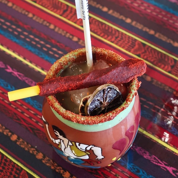 A colorful Mexican cocktail in a decorated mug with a chili-dusted straw and dried lime slices.