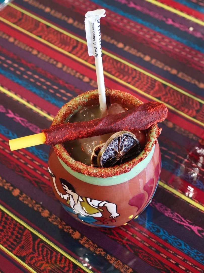 A colorful Mexican cocktail in a decorated mug with a chili-dusted straw and dried lime slices.
