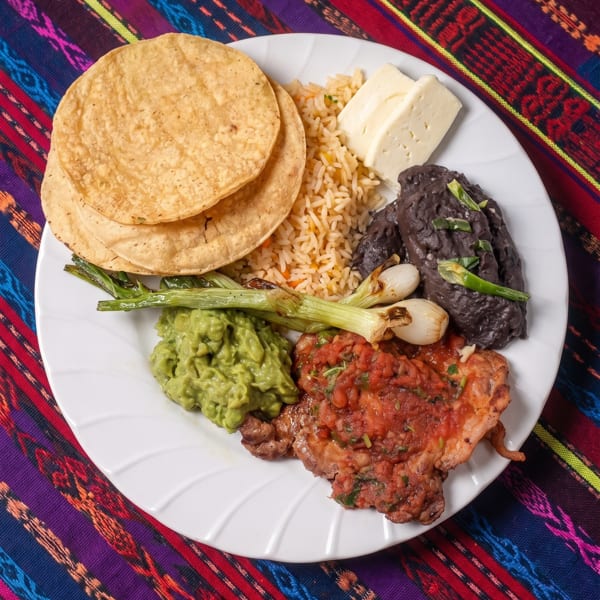 A white plate with steak, rice, black beans, guacamole, grilled onions, cheese, and tortillas.