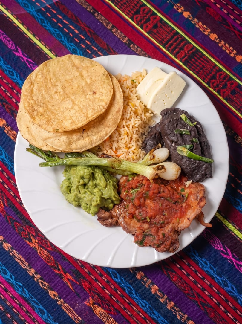 A white plate with steak, rice, black beans, guacamole, grilled onions, cheese, and tortillas.