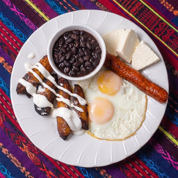 A white plate with fried plantains, black beans, fried eggs, sausage, and cheese on a colorful tablecloth.