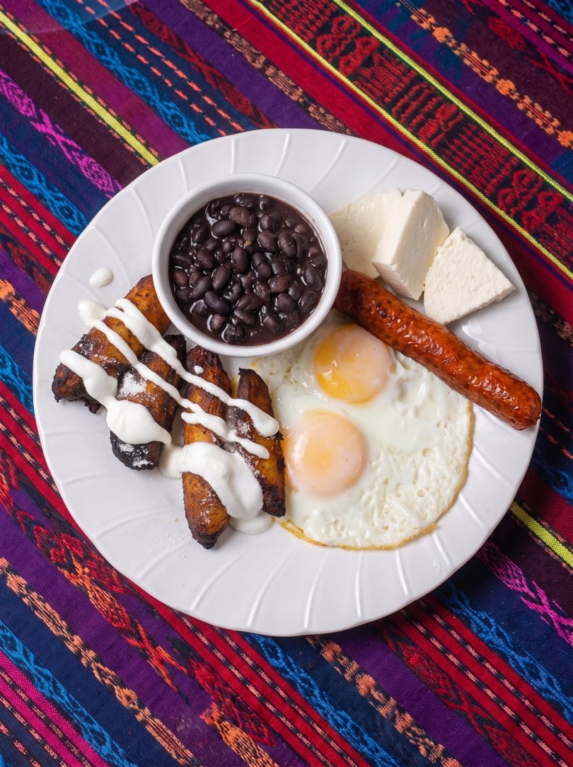 A white plate with fried plantains, black beans, fried eggs, sausage, and cheese on a colorful tablecloth.