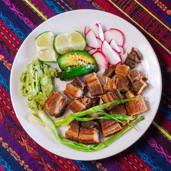 Crispy pork belly with guacamole, limes, radishes, jalapeños, and green onions on a colorful tablecloth.