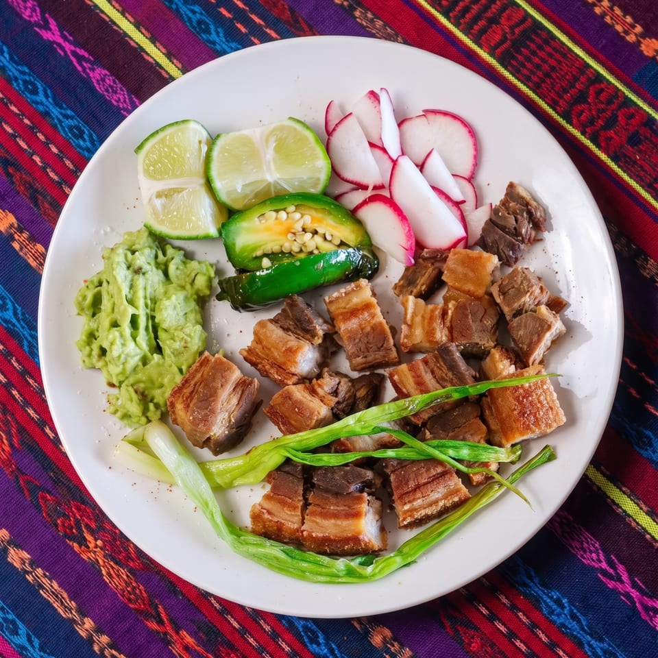Crispy pork belly with guacamole, limes, radishes, jalapeños, and green onions on a colorful tablecloth.