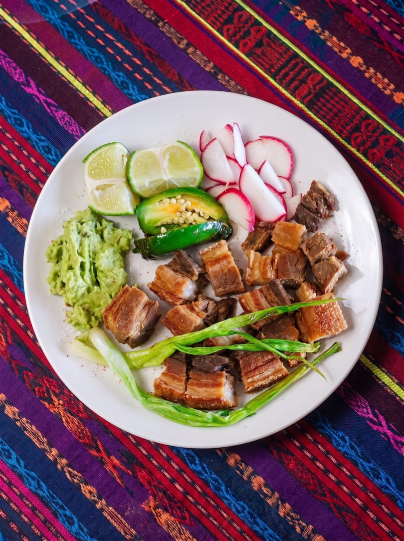 Crispy pork belly with guacamole, limes, radishes, jalapeños, and green onions on a colorful tablecloth.
