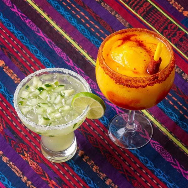 Two vibrant margaritas, one cucumber and one mango, on a colorful striped Mexican blanket.