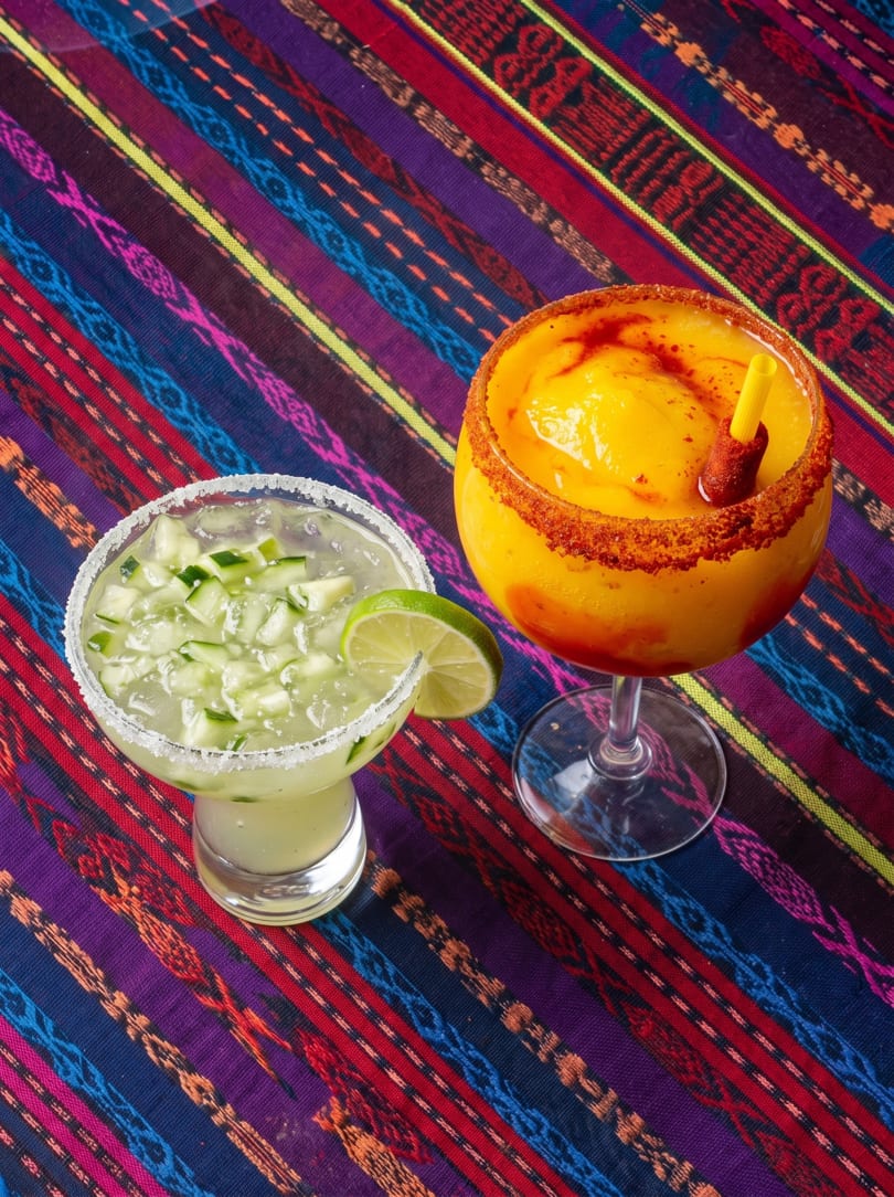Two vibrant margaritas, one cucumber and one mango, on a colorful striped Mexican blanket.