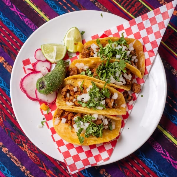 Three tacos with meat, onions, and cilantro, served with limes, radishes, and a green pepper.