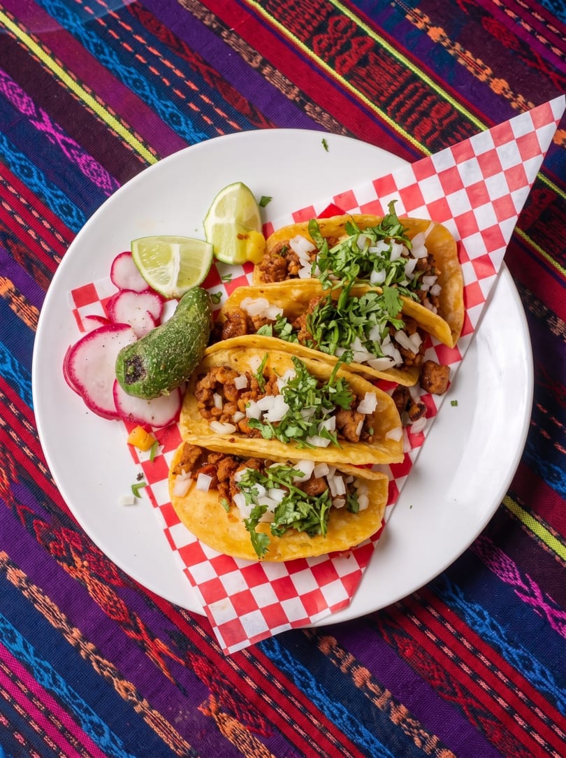 Three tacos with meat, onions, and cilantro, served with limes, radishes, and a green pepper.