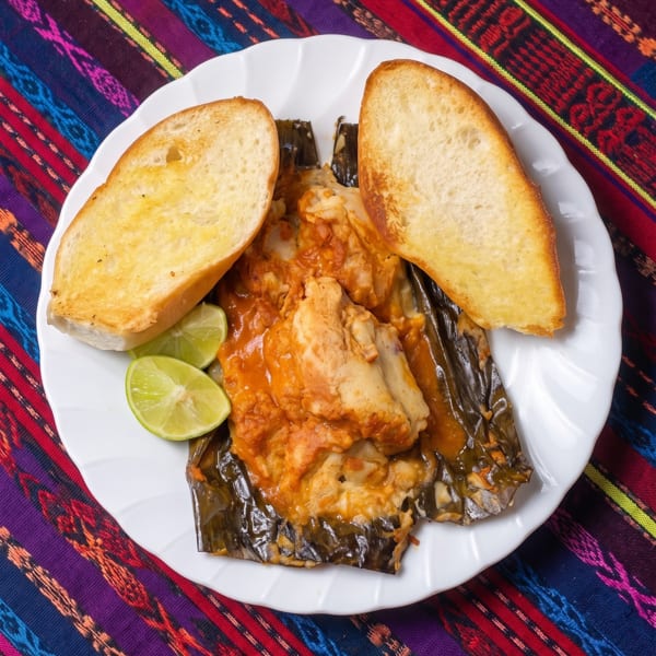 A white plate with a tamale in a banana leaf, two slices of bread, and lime wedges.