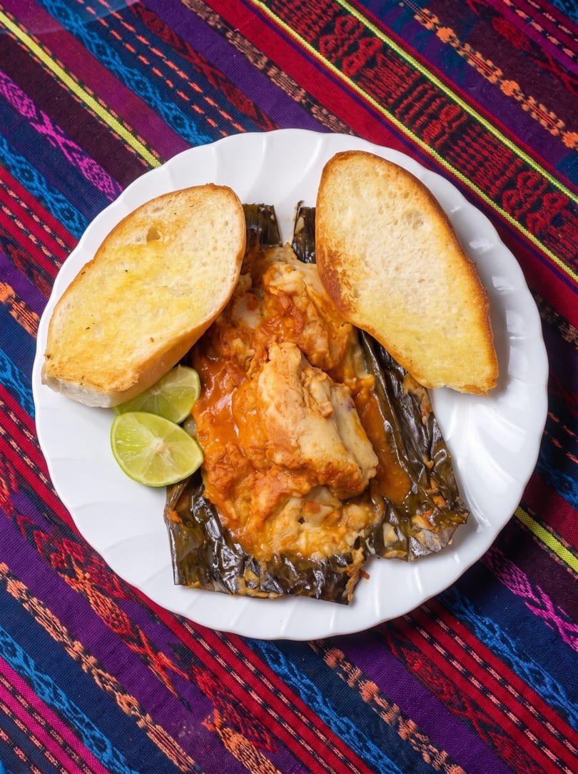 A white plate with a tamale in a banana leaf, two slices of bread, and lime wedges.